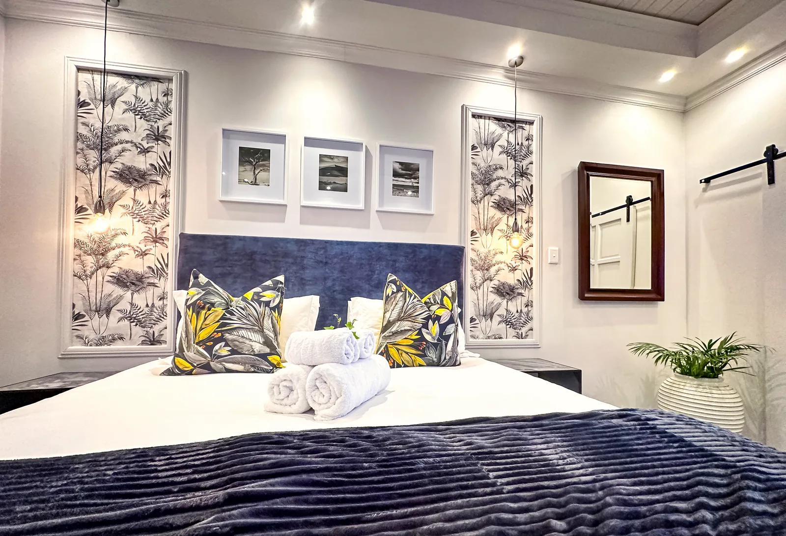 Made-up bed with rolled white towels, leaf-print cushions and a navy throw, framed by botanical wallpaper panels and three black-and-white prints